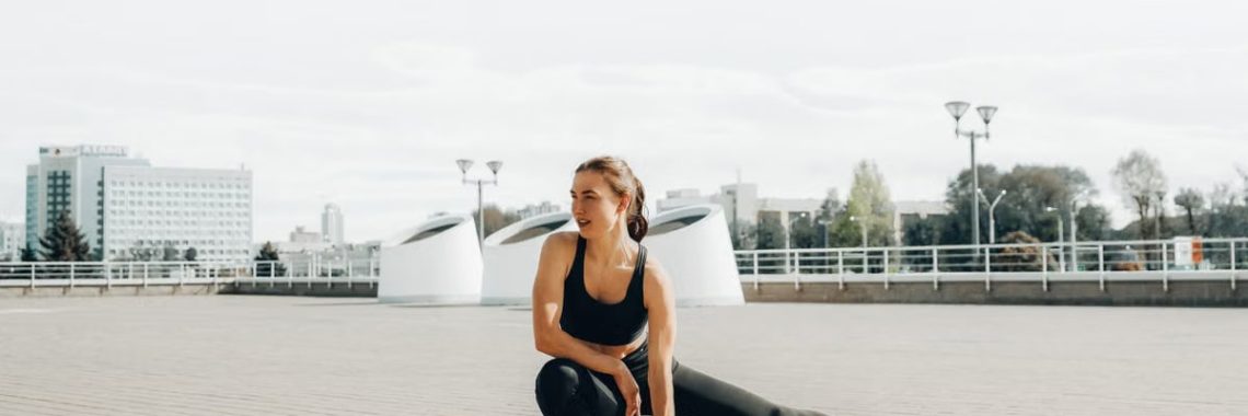 Young woman in black sportswear stretching outdoors in a sunny urban environment.